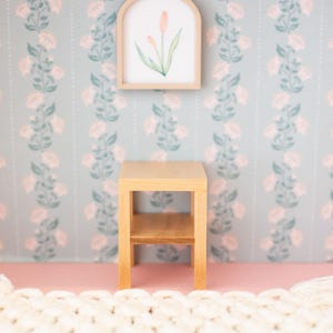 May include: A miniature wooden side table with a lower shelf stands on a pink surface. Above it, a framed watercolor painting of a flower is mounted on a floral wallpaper background. A cream-colored rug is in the foreground.