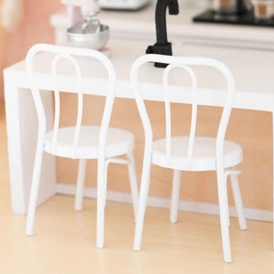 May include: Two white metal chairs with a curved back and a round seat. The chairs are placed in front of a white counter top in a dollhouse kitchen.