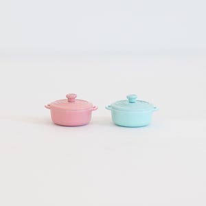 May include: Two small, pastel-coloured ceramic pots with lids. One is pink, the other is a light blue. Each pot has small handles and a knob on the lid. The pots are likely for cooking or serving small portions of food.