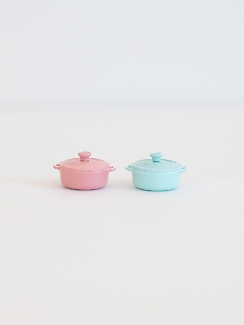 May include: Two small, pastel-coloured ceramic pots with lids. One is pink, the other is a light blue. Each pot has small handles and a knob on the lid. The pots are likely for cooking or serving small portions of food.