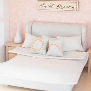 May include: A white bed with a gray headboard and a white duvet cover. There are three pillows on the bed, one with a sun design, one with a rainbow design, and one plain white pillow. The bed is in a room with pink floral wallpaper and a sign above the headboard that says "Sweet Dreams".
