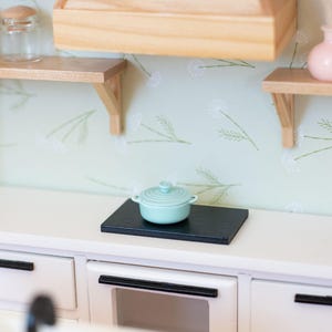 May include: A miniature kitchen scene with a light blue lidded pot on a black surface. The kitchen features white cabinets, a wooden range hood, and shelves. The walls are decorated with a floral pattern.