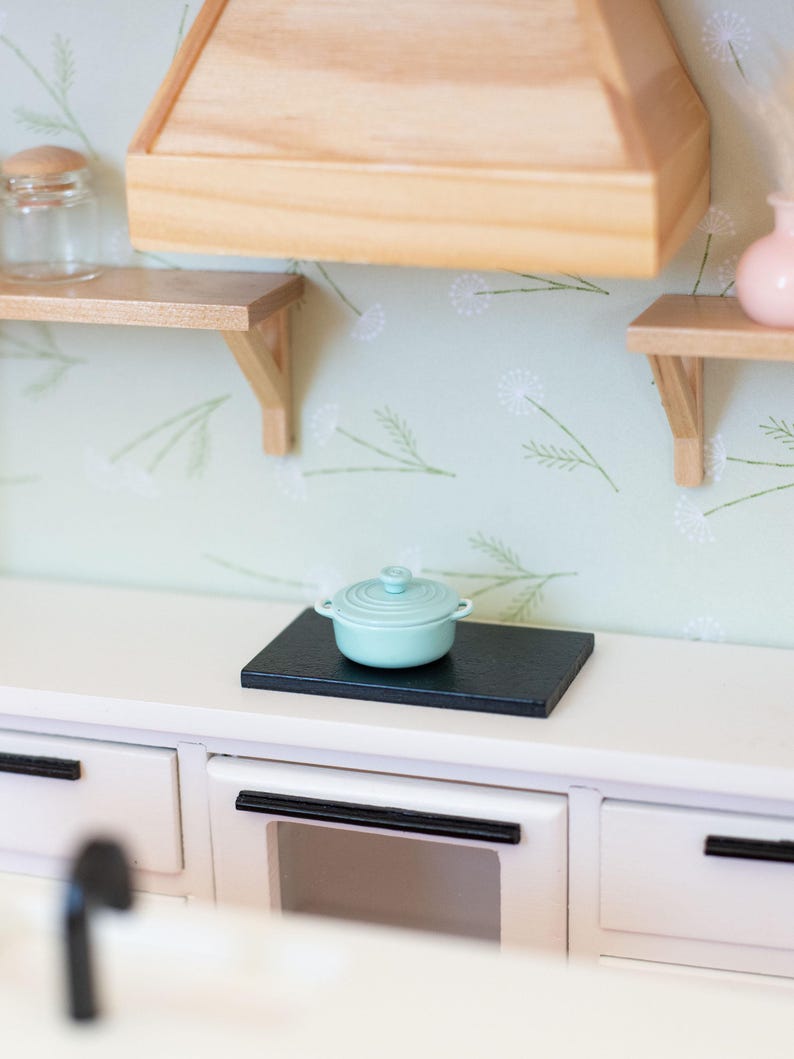 May include: A miniature kitchen scene with a light blue lidded pot on a black surface. The kitchen features white cabinets, a wooden range hood, and shelves. The walls are decorated with a floral pattern.