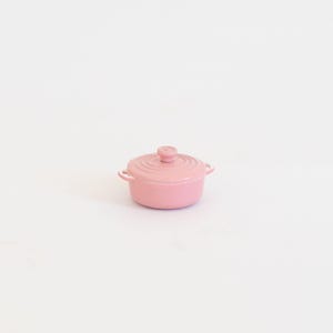 May include: A small, pink lidded pot with two side handles and a round knob on the lid. The pot is made of a solid material and has a smooth, glossy finish. The pot is centered on a white background.