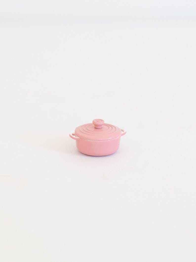 May include: A small, pink lidded pot with two side handles and a round knob on the lid. The pot is made of a solid material and has a smooth, glossy finish. The pot is centered on a white background.