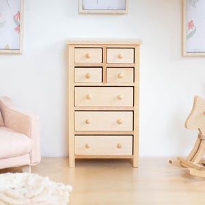 May include: A miniature wooden dresser with seven drawers, featuring small round knobs. The dresser is light-colored and stands on a wooden floor. Two framed floral prints hang on the white wall behind it.