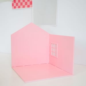 May include: A pink miniature house with a pitched roof and a window. The house is made of a solid material and has a simple, clean design. The background features framed artwork in pink, gray, and green.