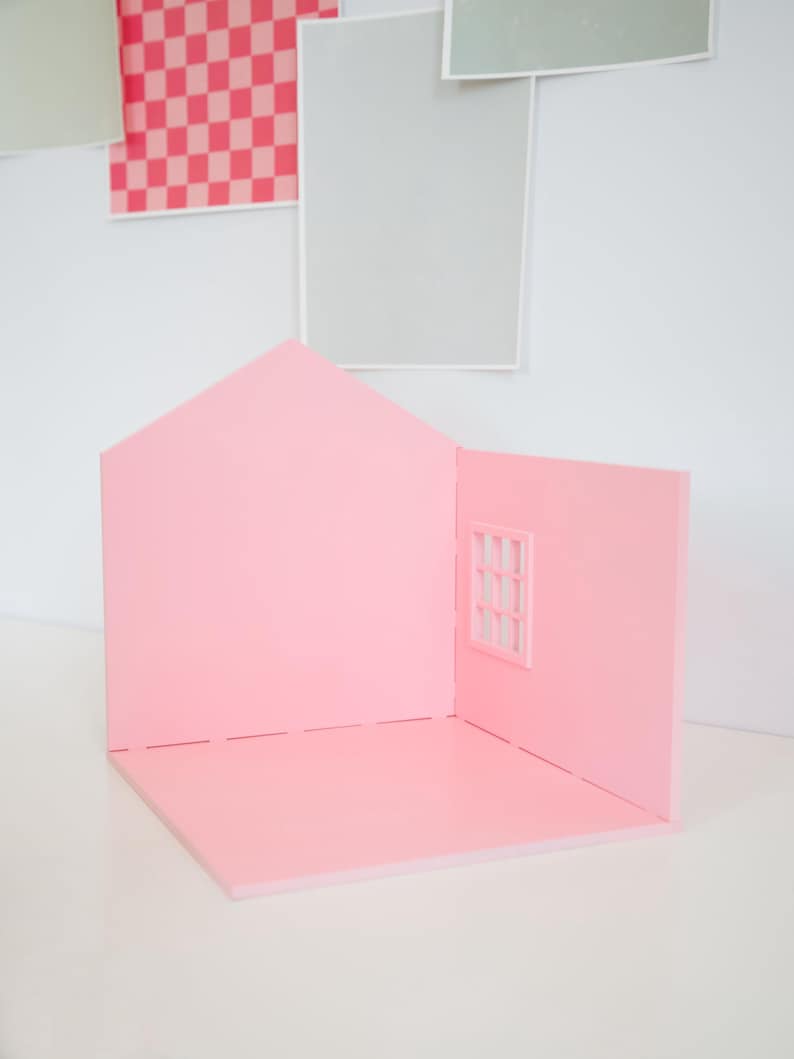 May include: A pink miniature house with a pitched roof and a window. The house is made of a solid material and has a simple, clean design. The background features framed artwork in pink, gray, and green.