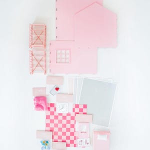 May include: A disassembled pink dollhouse kit with various components, including walls, a roof, and furniture. The kit includes a checkered floor, a clock, and small accessories. The pieces are arranged on a white surface.