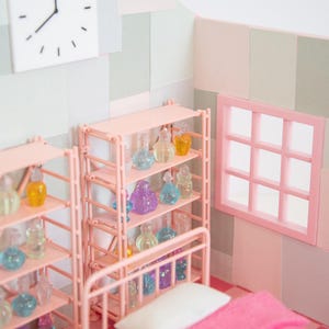 May include: A miniature bedroom scene with pink furniture, including a bed with a pink blanket and a shelf unit filled with colorful glass bottles. A white clock hangs on the wall. The room has a window and is decorated in pastel colors.