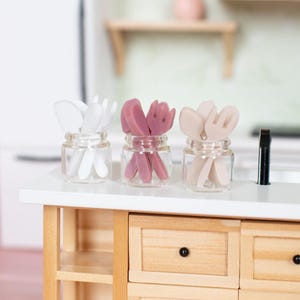 May include: Three small glass jars filled with miniature utensils in white, mauve, and beige. The jars are arranged on a white countertop in a miniature kitchen setting. The kitchen features light wood cabinetry and a white refrigerator.
