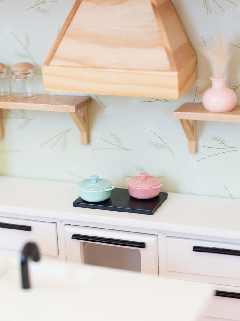 May include: Miniature kitchen scene with a white countertop, featuring a black cooktop holding a light blue and a pink lidded pot. A wooden range hood and shelves with a vase and jars are visible.