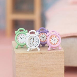 May include: Four miniature alarm clocks in pastel colors: green, white, purple, and pink. Each clock has a round face with black hands and a small stand. The clocks are displayed on a wooden block.