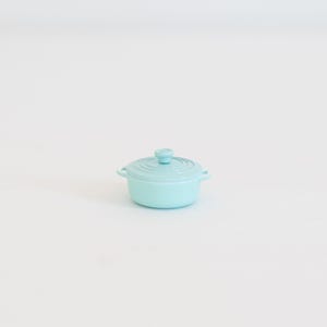 May include: A small, pale blue cast iron casserole dish with a matching lid. The pot has two side handles and a small knob on the lid. The cookware is designed for stovetop or oven use.