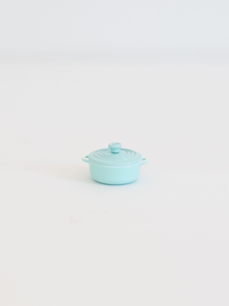 May include: A small, pale blue cast iron casserole dish with a matching lid. The pot has two side handles and a small knob on the lid. The cookware is designed for stovetop or oven use.