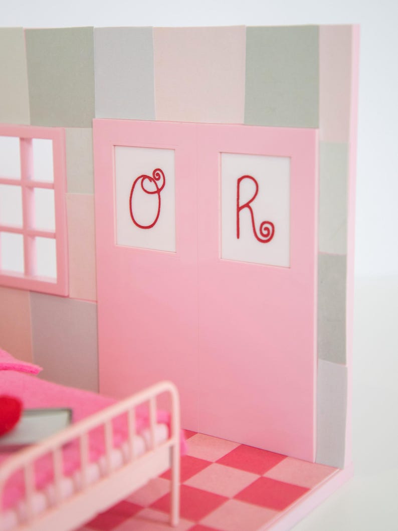 May include: A miniature pink bedroom scene with a bed, window, and a door with the letters "O" and "R" in red. The floor has a pink and red checkered pattern, and the walls are a mix of pink and gray.