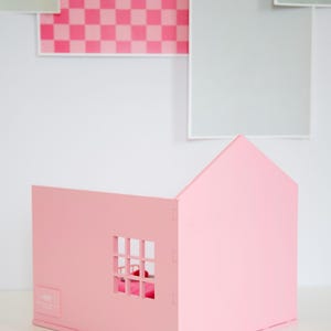 May include: A pink wooden dollhouse with a pitched roof and a window. The dollhouse is made of interlocking pieces and sits on a white surface. The background features pink and gray squares.