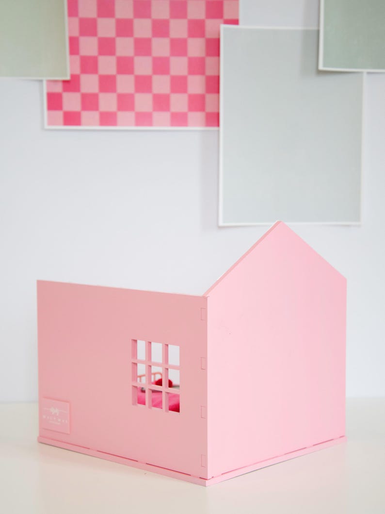May include: A pink wooden dollhouse with a pitched roof and a window. The dollhouse is made of interlocking pieces and sits on a white surface. The background features pink and gray squares.
