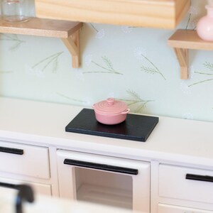 May include: A miniature kitchen scene with a pink lidded pot on a black surface, atop a white cabinet. The kitchen features wooden shelves, light green floral wallpaper, and a wooden range hood.