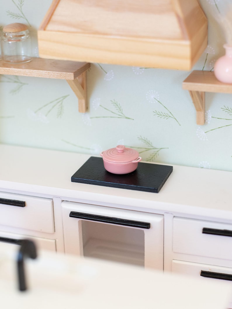 May include: A miniature kitchen scene with a pink lidded pot on a black surface, atop a white cabinet. The kitchen features wooden shelves, light green floral wallpaper, and a wooden range hood.