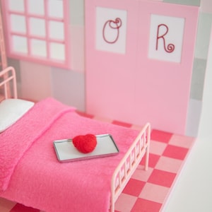 May include: A miniature pink bedroom scene with a bed, window, and door. A red heart sits on a tray on the bed. The bed has a pink blanket and a white pillow. The floor is a pink and red checkerboard pattern.