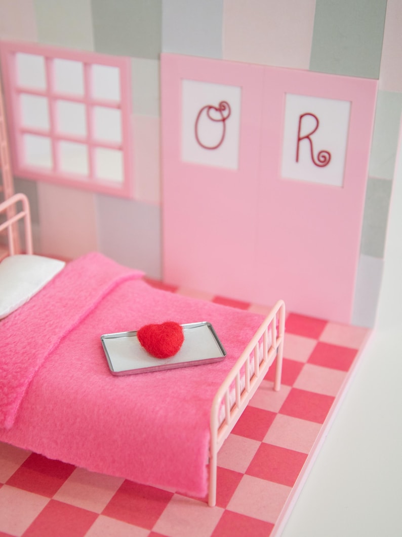 May include: A miniature pink bedroom scene with a bed, window, and door. A red heart sits on a tray on the bed. The bed has a pink blanket and a white pillow. The floor is a pink and red checkerboard pattern.