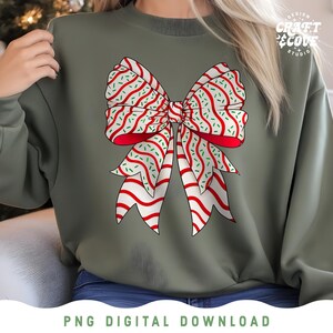 May include: An olive green sweatshirt featuring a large, festive bow graphic. The bow has red and white striped ribbons with green sprinkles. The design is a PNG digital download. The text "Design Craft & Cove Studio" is visible.