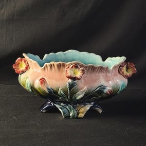 Pode incluir: Uma taça de cerâmica com rebordo recortado e interior azul claro. O exterior apresenta um gradiente de rosa para azul escuro, decorado com motivos florais tridimensionais em relevo nas cores vermelho, amarelo e verde, sobre três pés.