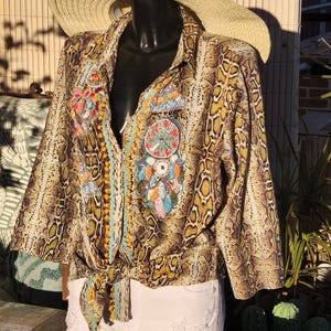 Peut inclure: Une chemise à imprimé peau de serpent avec des détails brodés colorés et un devant noué. La chemise est associée à un short en jean blanc et un chapeau de paille à larges bords. La tenue est présentée sur un mannequin.