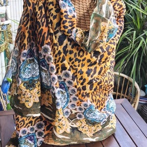 May include: A patterned scarf with a leopard print design in shades of yellow, orange, and black. The scarf also features floral and paisley patterns in blue, white, and green. A straw hat sits atop a wicker form.