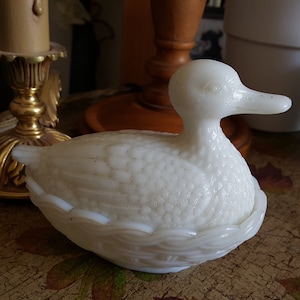 Pode incluir: Um prato de vidro de leite branco em forma de pato com um design de ninho. O pato está sentado em uma mesa com uma superfície com padrão marrom e branco.