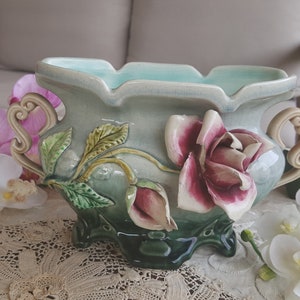 Peut inclure: Un pot en céramique avec un glaçage bleu clair et des détails floraux complexes. Le pot présente un motif en relief de roses et de feuilles, avec un jeu de couleurs vert et marron. Le pot a deux poignées sur les côtés.
