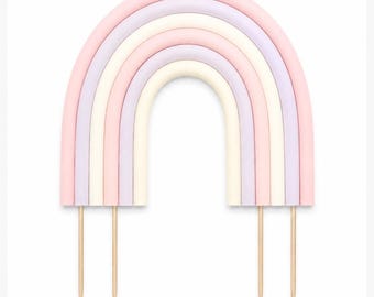Puede incluir: Un adorno para pastel en forma de arcoíris con cinco bandas arqueadas en colores pastel alternos rosa, lavanda y crema. El arco está sostenido por cuatro palitos de madera finos y puntiagudos que sobresalen de la base.