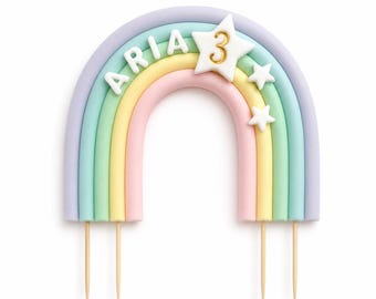 May include: A pastel-colored rainbow cake topper featuring the name ARIA in white letters and a white star with the gold number 3. The rainbow has bands of purple, teal, yellow, and pink, with two additional small white stars and wooden picks.