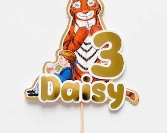 Puede incluir: Un adorno para tarta con un tigre y un niño bebiendo con pajitas, que incluye un gran número 3 dorado y el nombre Daisy en letras doradas. El diseño está montado sobre un fino palito de madera y presenta una ilustración con detalles dorados.