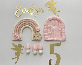 May include: A pink and brown rainbow, a pink fairy door, three pink and white mushrooms, a gold fairy silhouette, and the name "Evelyn" and the number "5" in gold glitter.