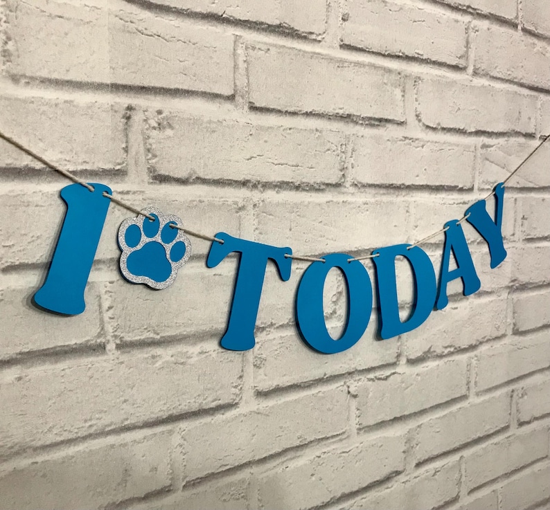 Puede incluir: Una pancarta azul que deletrea las palabras I TODAY con un icono de huella de pata con purpurina plateada entre la letra I y la palabra TODAY. Las letras est&aacute;n ensartadas en un cordel fino, creando un letrero colgante decorativo.