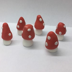 May include: A group of seven miniature mushroom ornaments. Each mushroom features a vibrant red cap adorned with white spots and a white stem. The ornaments are set on a white surface.