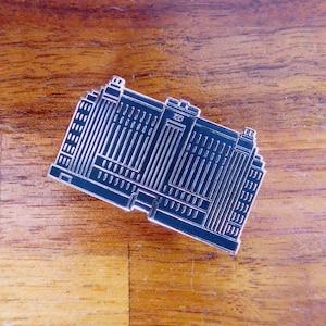 May include: Black metal pin shaped like a building with many windows. The pin is a stylized representation of a skyscraper.