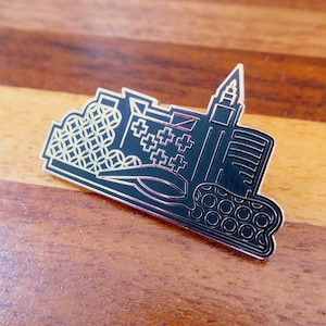 May include: Black and gold enamel pin featuring a stylised cityscape with a tall building, a building with a cross pattern, and a building with a circular pattern.