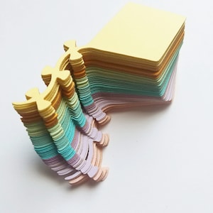 May include: A stack of pastel colored paper notes with a unique, decorative edge. The top note is yellow, and the stack fades to light blue, then pink.