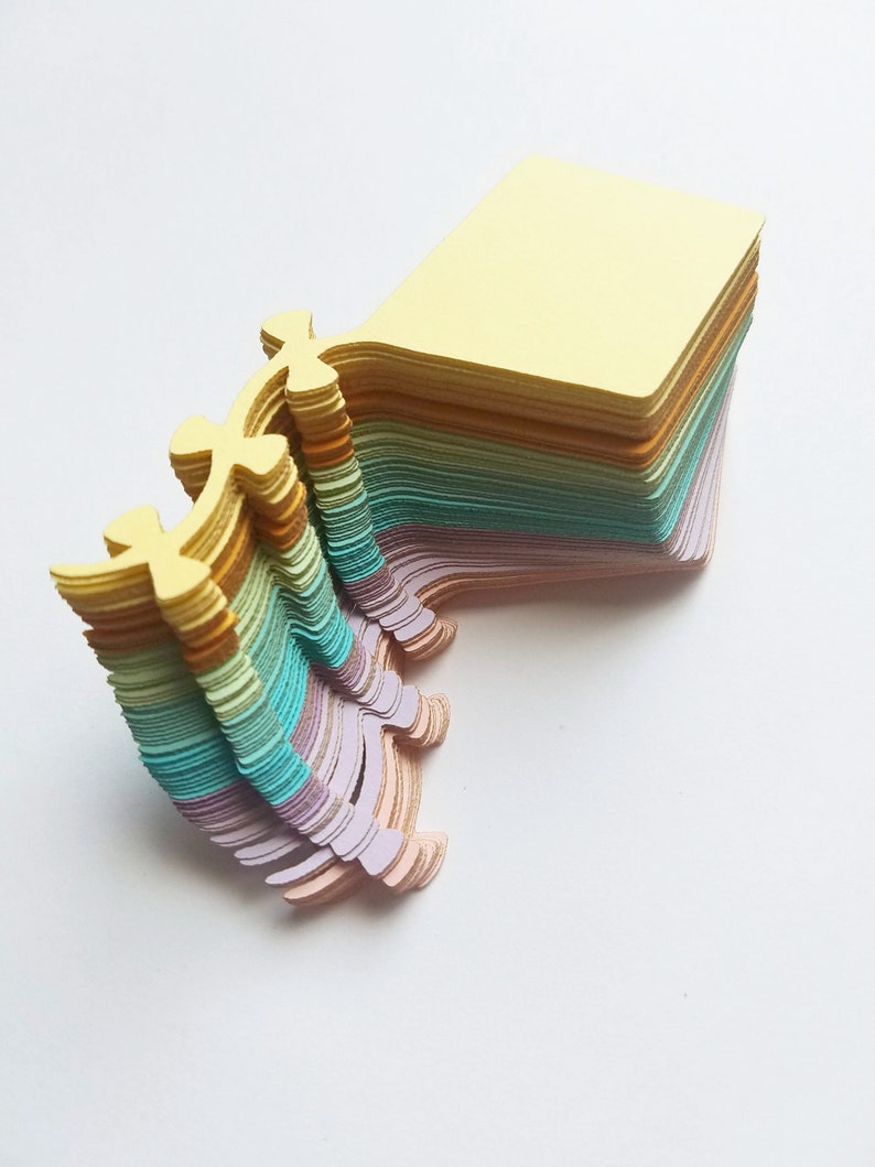 May include: A stack of pastel colored paper notes with a unique, decorative edge. The top note is yellow, and the stack fades to light blue, then pink.