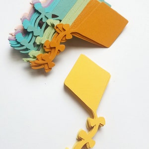 May include: A stack of colorful paper cutouts in the shape of kites. The kites are layered in shades of purple, blue, green, yellow, and orange. The kites have a decorative design on the bottom.