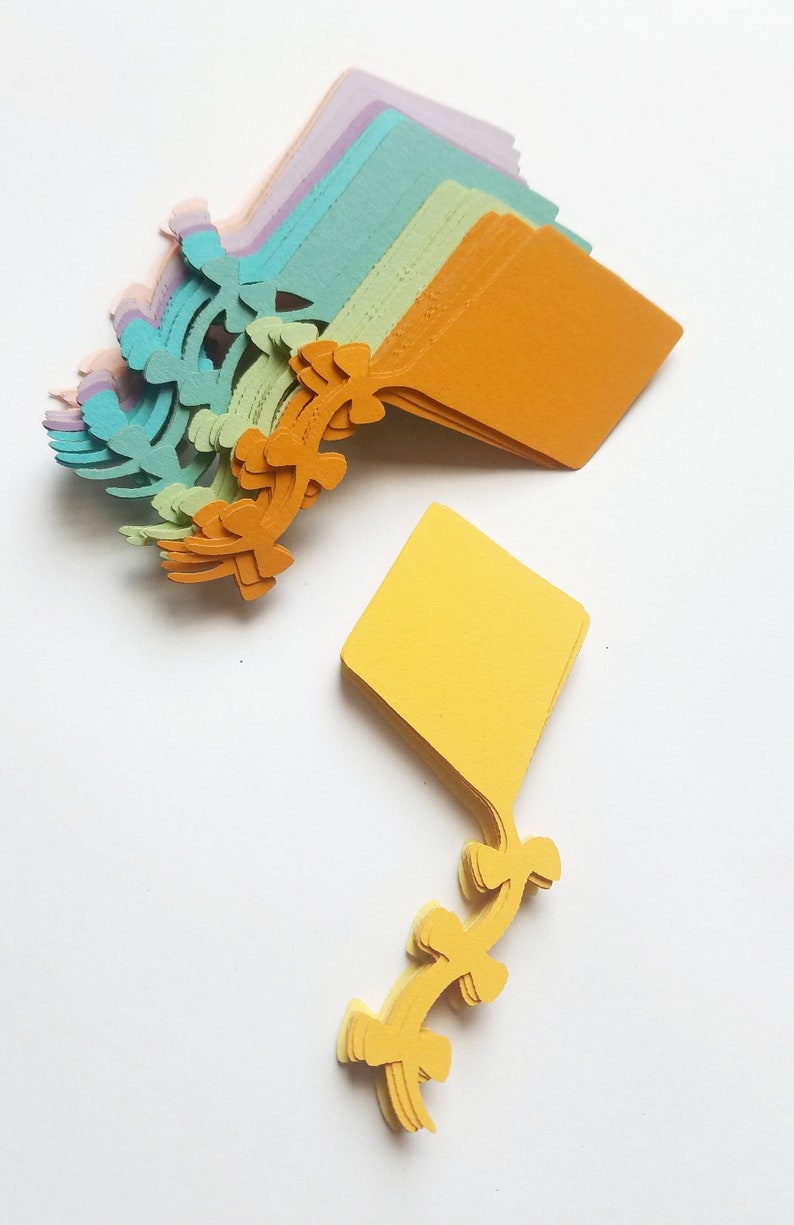 May include: A stack of colorful paper cutouts in the shape of kites. The kites are layered in shades of purple, blue, green, yellow, and orange. The kites have a decorative design on the bottom.