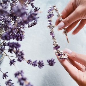 May include: A silver headband with purple crystal accents. The headband is held in a hand.