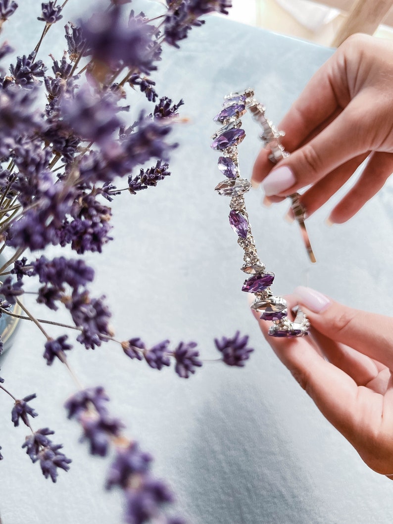 May include: A silver headband with purple crystal accents. The headband is held in a hand.