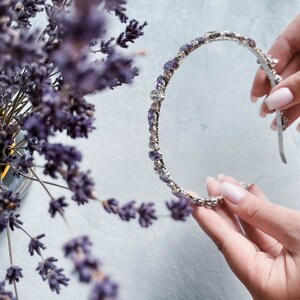 May include: A silver headband with purple rhinestones. The headband is held in a hand.
