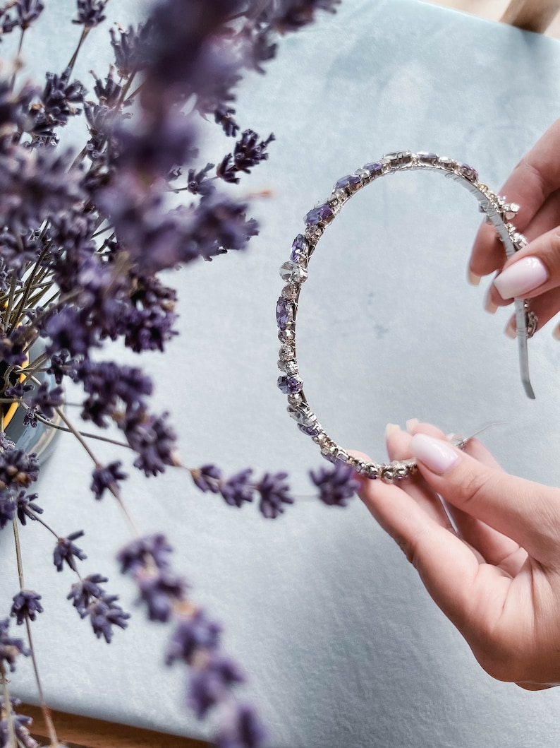 May include: A silver headband with purple rhinestones. The headband is held in a hand.