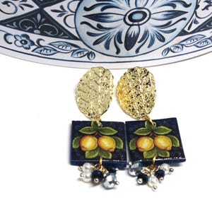 Op de afbeelding: Goudkleurige oorbellen met getextureerde bladvormige tops en vierkante, marineblauwe hangers. Elke hanger heeft een schilderij van twee gele citroenen met groene bladeren. Kleine parel- en kralenclusters hangen eronder.