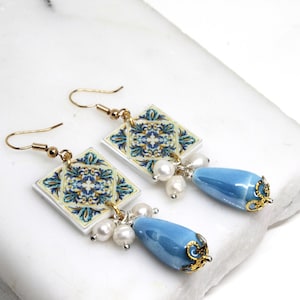 Peut inclure: Boucles d'oreilles pendantes composées de carreaux de céramique carrés avec un motif floral bleu et jaune. Chaque boucle d'oreille comporte des perles blanches et une perle en forme de goutte d'eau bleu clair avec des accents dorés.